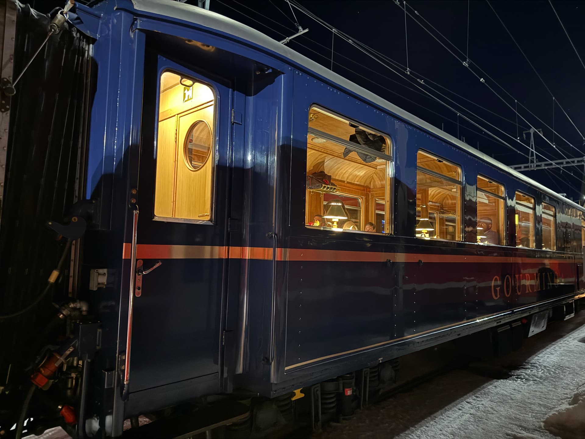 The blue Gourmino dining carriage at night, warm windows above the snowy platform