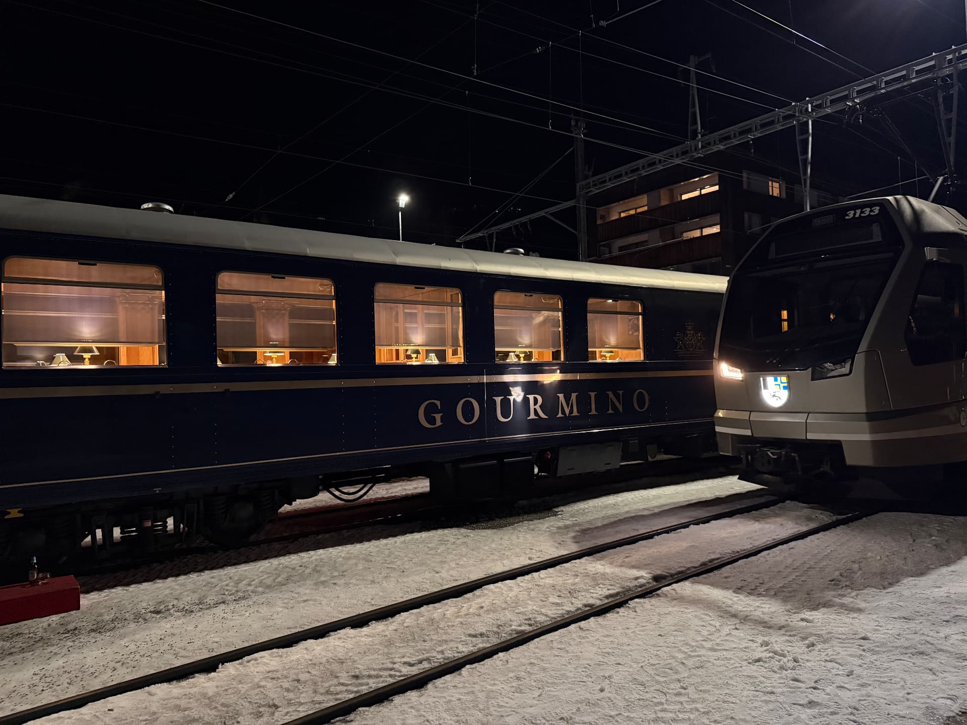 The lit Gourmino carriage at night beside a modern Rhaetian Railway train at Davos Platz