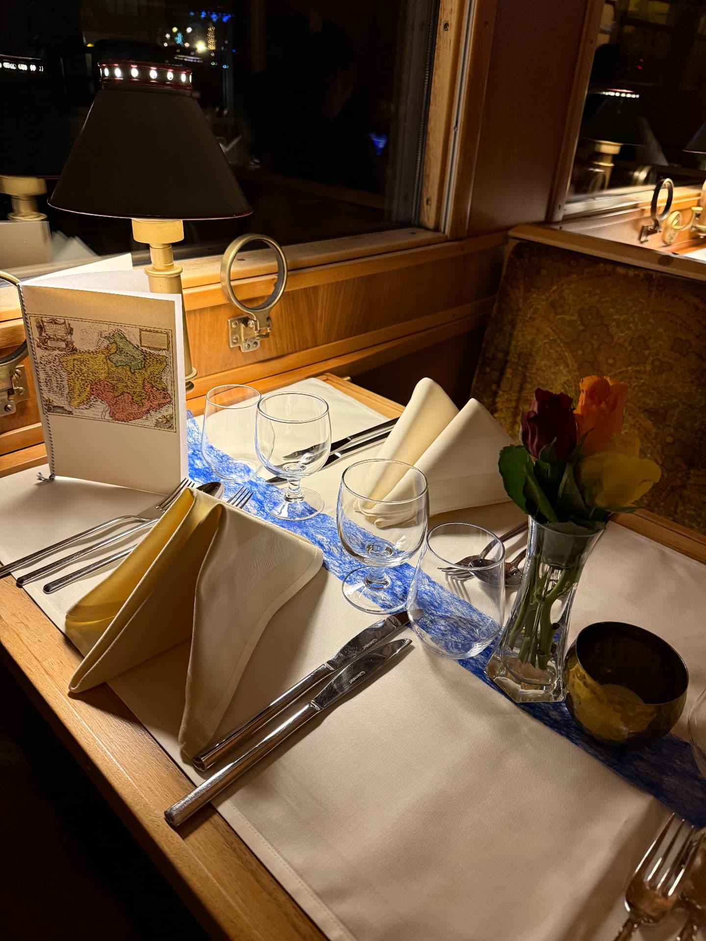 Place setting with an illustrated menu card, brass lamp and roses beside the carriage window